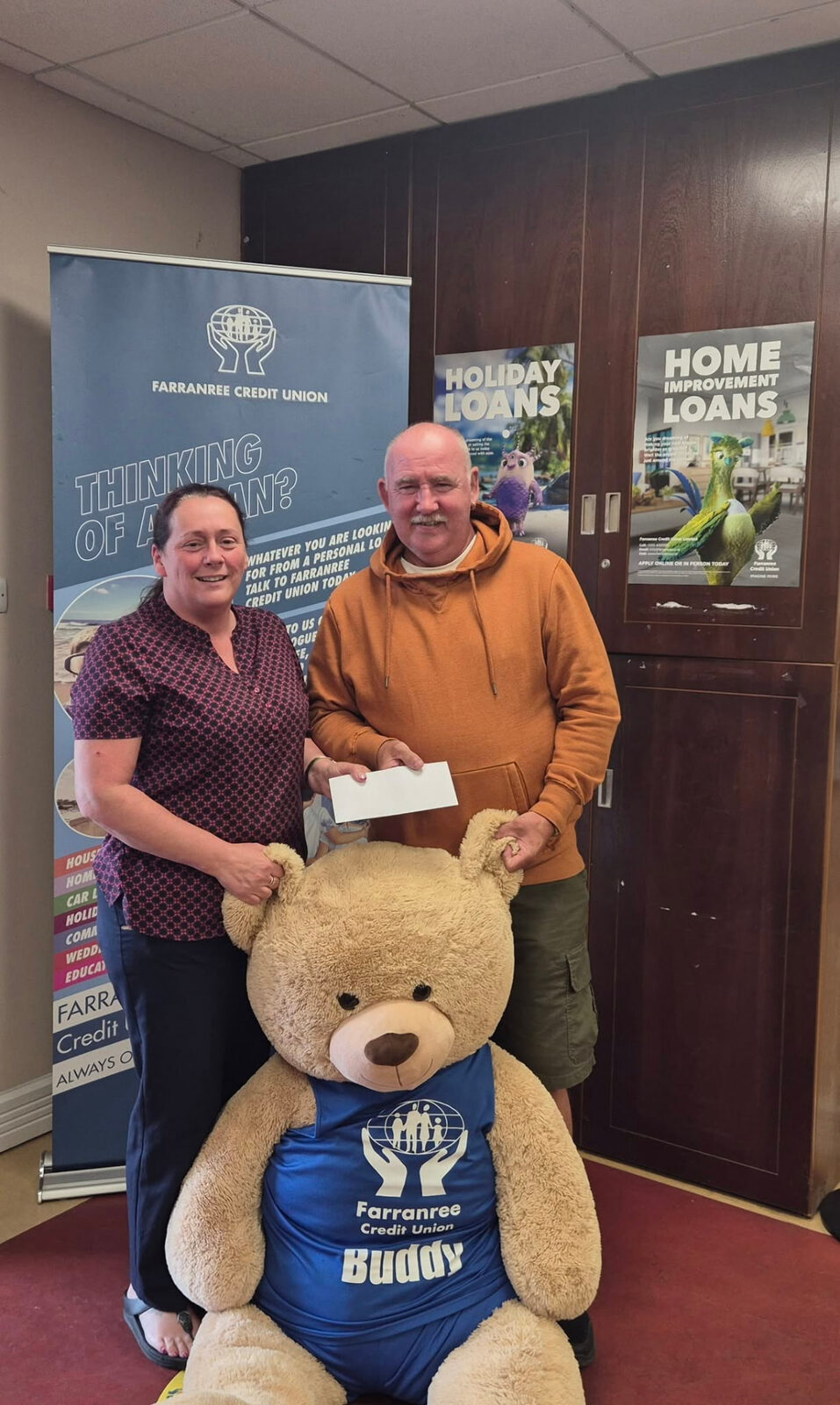 Two people holding an envelope beside a large teddy bear
